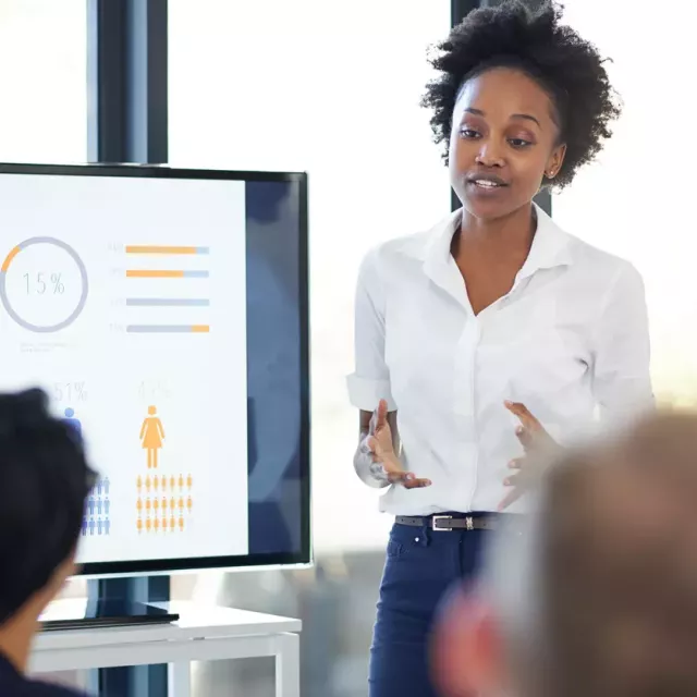 A business woman presents in front of an audience with a screen showing charts and metrics next to her.