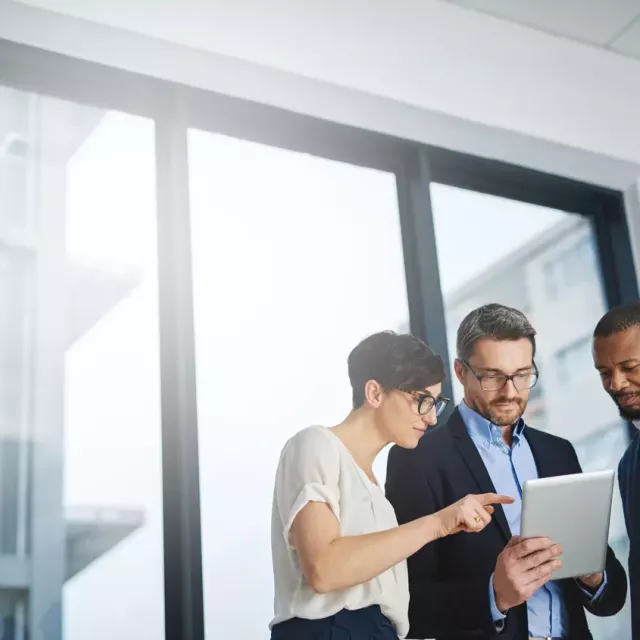 Three diverse coworkers in professional attire look at a tablet computer together while standing in front of tall transparent glass office windows with similar tall buildings visible outside in the daylight.