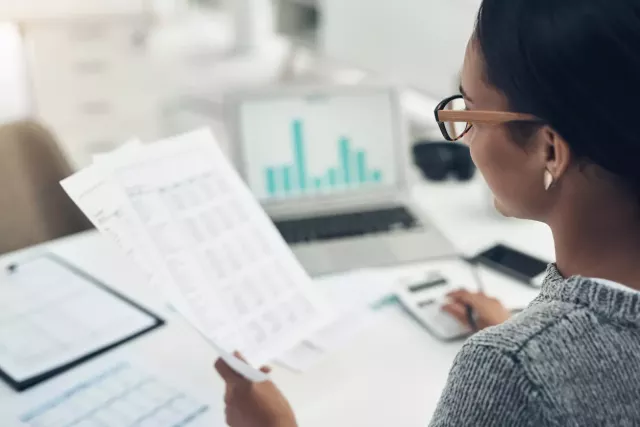 Woman sitting at a desk holding printed spreadsheets, using a calculator.