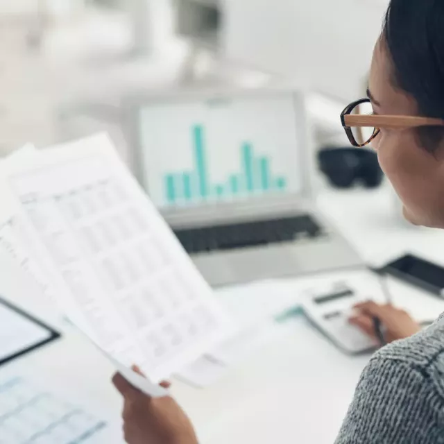 Woman sitting at a desk holding printed spreadsheets, using a calculator.