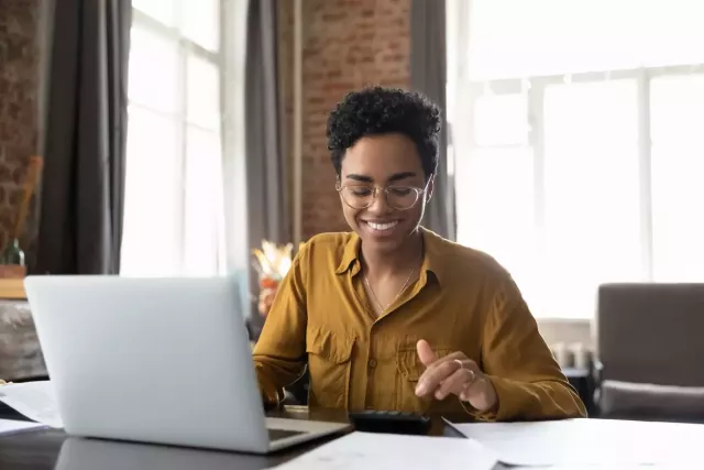 Young black woman wearing glasses and a gold shirt sits at a desk with laptop in a brightly lit room with tall windows and exposed brick walls.