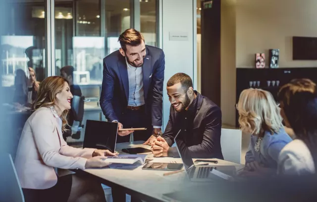 A group of diverse professionals review revenue figures at a table with printed graphs and laptops while smiling.