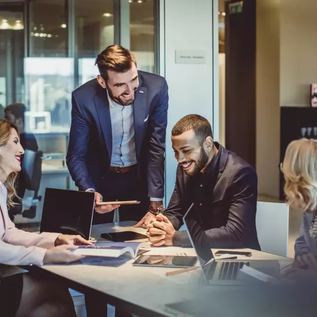 A group of diverse professionals review revenue figures at a table with printed graphs and laptops while smiling.