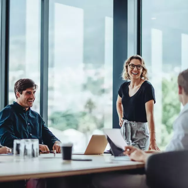 Woman in business casual clothes with glasses leans one hand on a conference table and laughs with coworkers in an office with large glass windows.