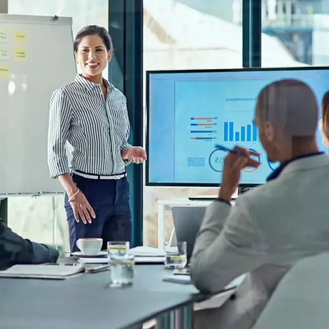 A woman wearing business casual presents sales KPIs on a digital screen in a bright modern conference room.