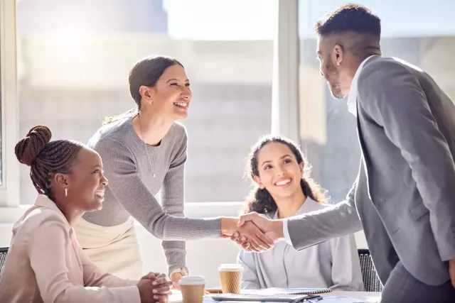 Young professionals shake hands over a conference table in an office with large glass windows.