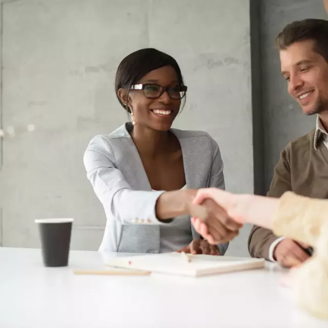 Smiling businesswoman shakes hands with female colleague  over a conference table, while male colleagues smiles in the background.