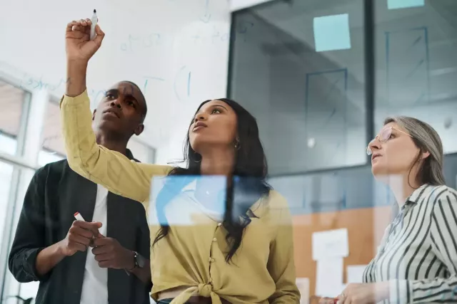 Young professional draws lines on a clear board or window, photographed from the other side, with coworkers beside her.