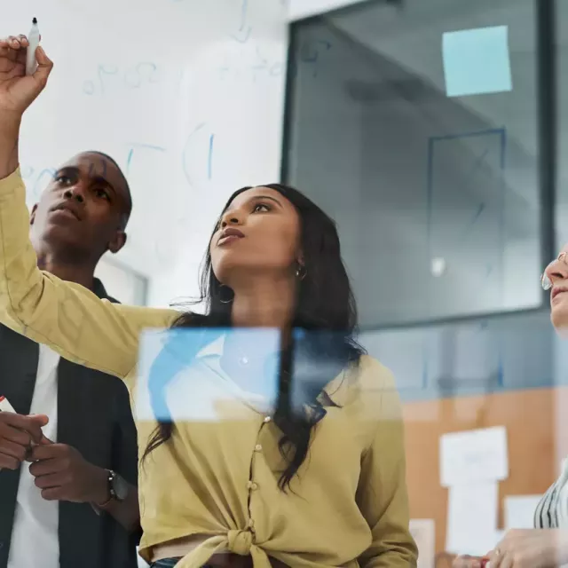 Young professional draws lines on a clear board or window, photographed from the other side, with coworkers beside her.