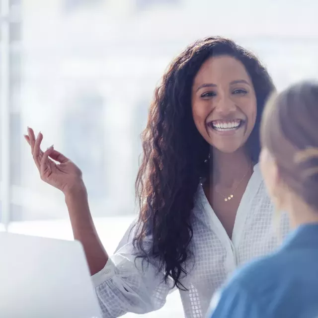 Two woman site at a table in a bright office with large glass windows. One is smiling toward the camera at the other.