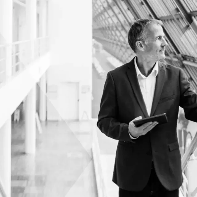 Man in suit jacket look out of large glass windows in a modern office while holding a tablet.