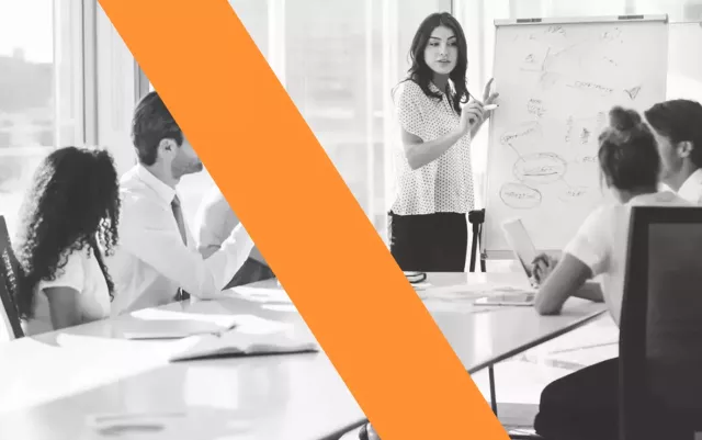 A black and white photo of a woman standing at chart turned and facing a group of people sitting at a conference table blog header for Xactly.