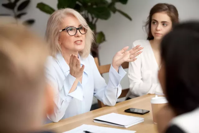 A business leader gestures while sitting at a conference table, speaking to colleagues.