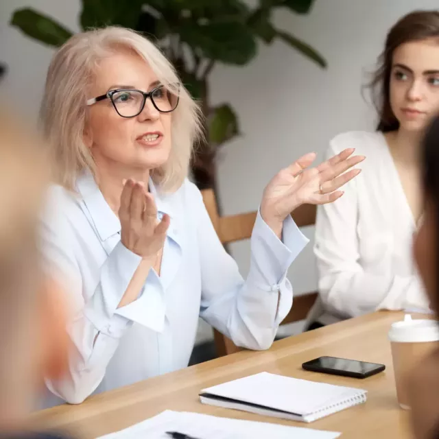 A business leader gestures while sitting at a conference table, speaking to colleagues.