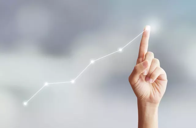 Hand with index finger extended drawing upward curve line and point chart in light with clouds in the background representing revenue growth.