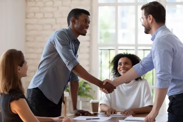 Two men in business casual clothes shake hands over a conference table.