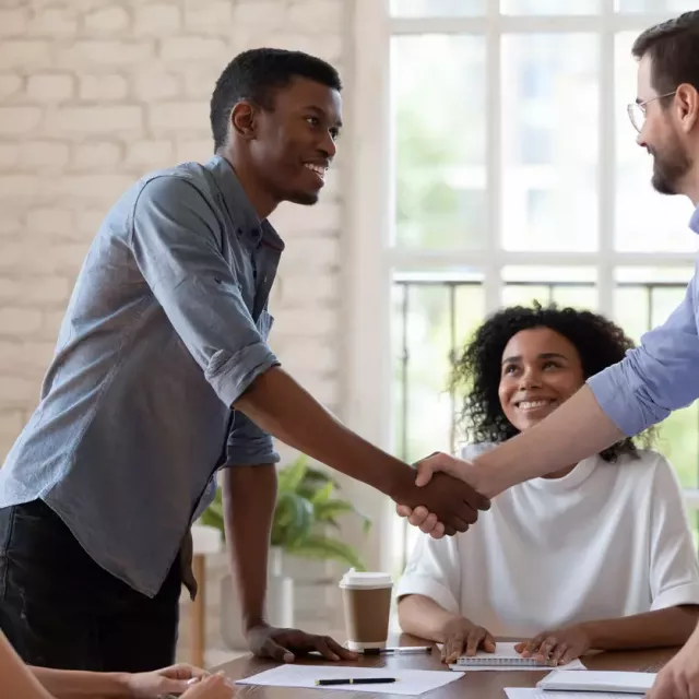 Two men in business casual clothes shake hands over a conference table.