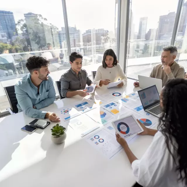 A group wearing business casual clothing sit around a white conference table discussing budget planning documents on which graphs are visible. In the large glass windows behind them, a cityscape is visible.