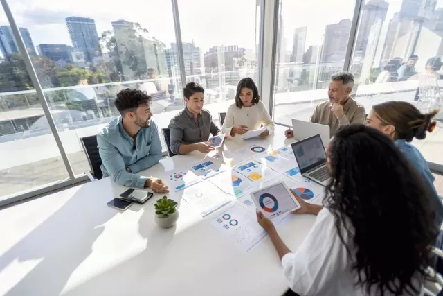 A group wearing business casual clothing sit around a white conference table discussing budget planning documents on which graphs are visible. In the large glass windows behind them, a cityscape is visible.